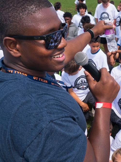Kelvin wearing sunglasses and a UT Tyler lanyard speaking into a microphone while holding a water bottle, surrounded by children in white event t-shirts outdoors.