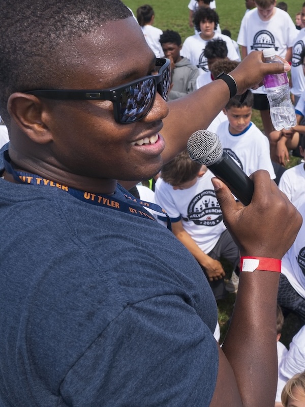 Kelvin wearing sunglasses and a UT Tyler lanyard speaking into a microphone while holding a water bottle, surrounded by children in white event t-shirts outdoors.