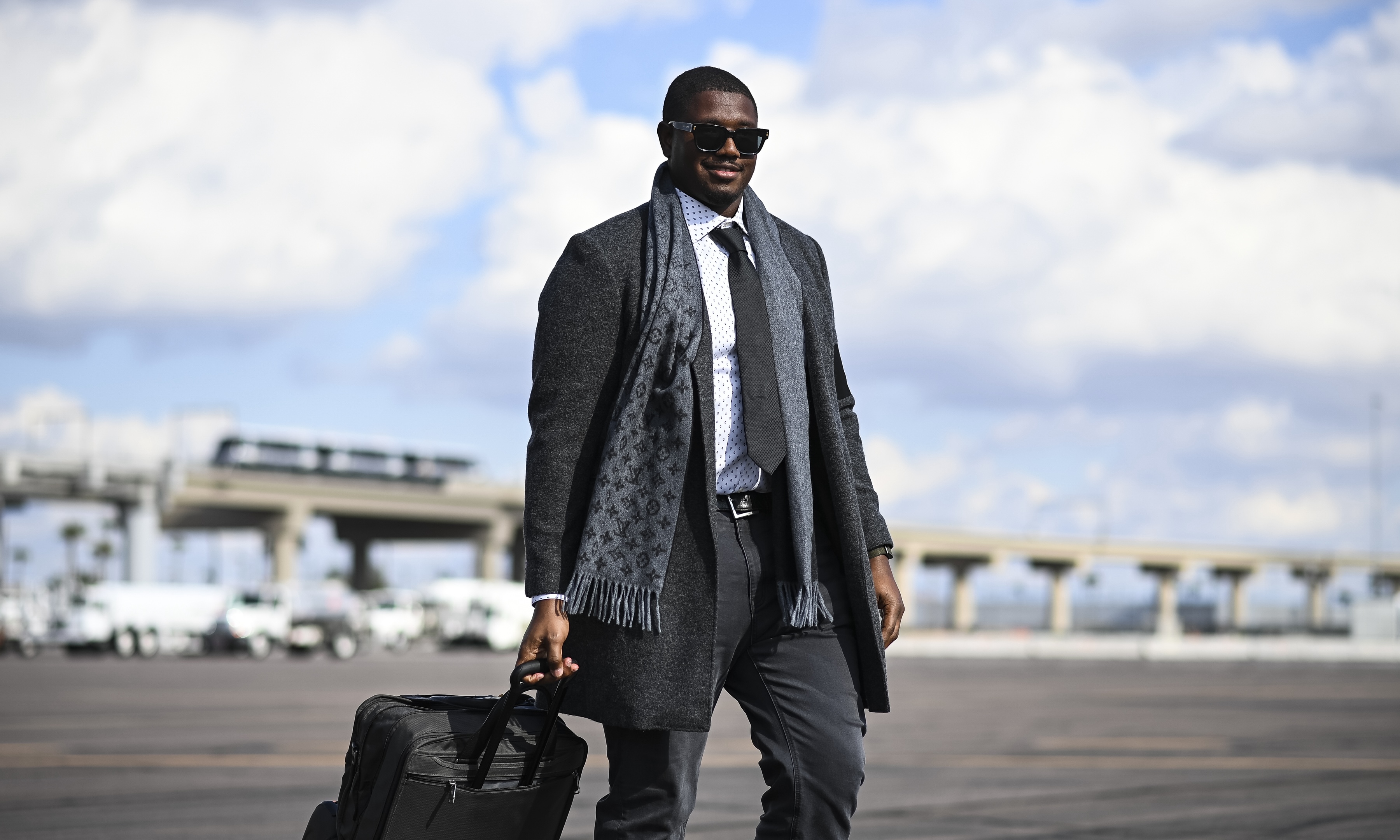 Kelvin in sunglasses, a gray coat, patterned scarf, and tie walking on a tarmac while pulling a black rolling suitcase.