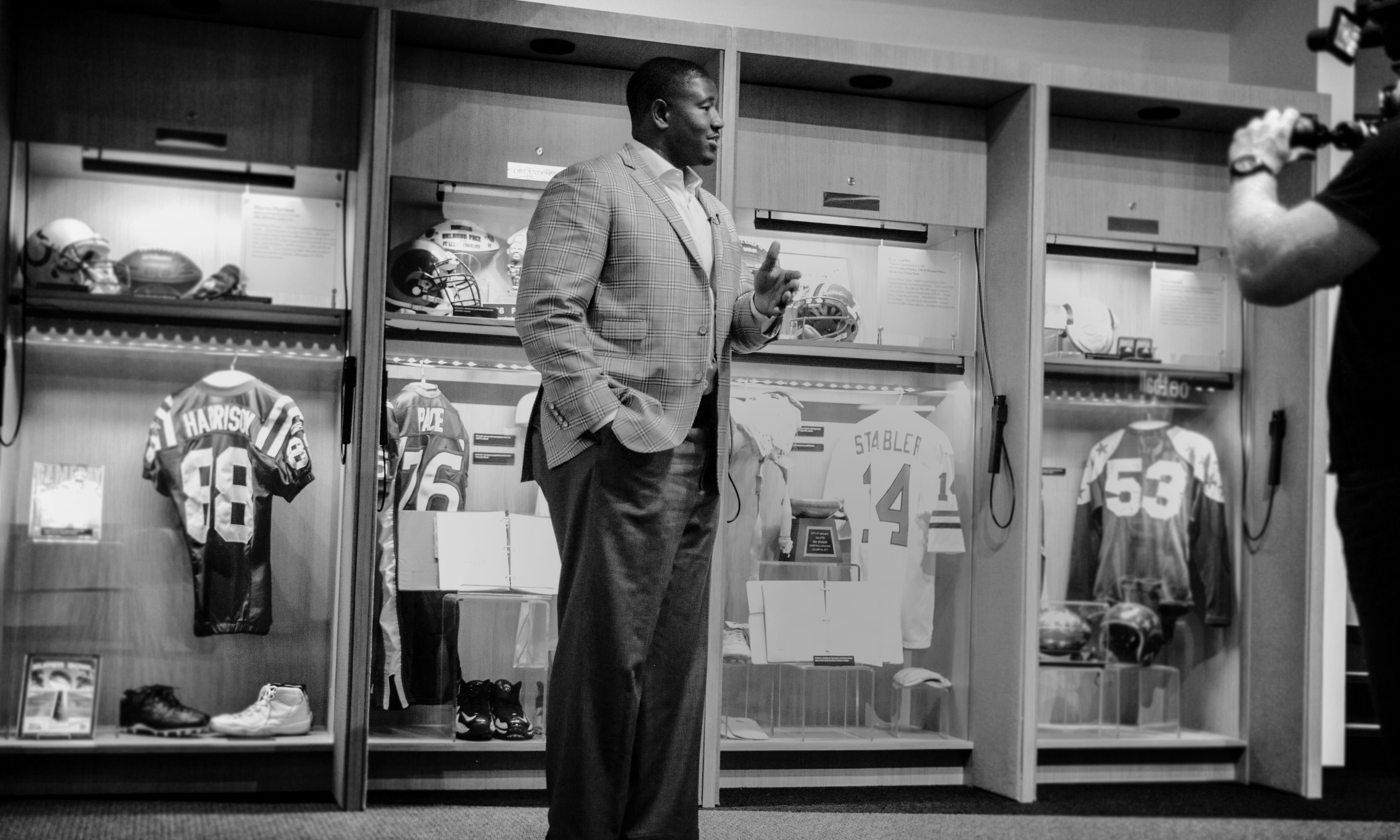 Kelvin in a plaid blazer speaking in front of a wall of illuminated sports memorabilia displays including football jerseys, helmets, and shoes.