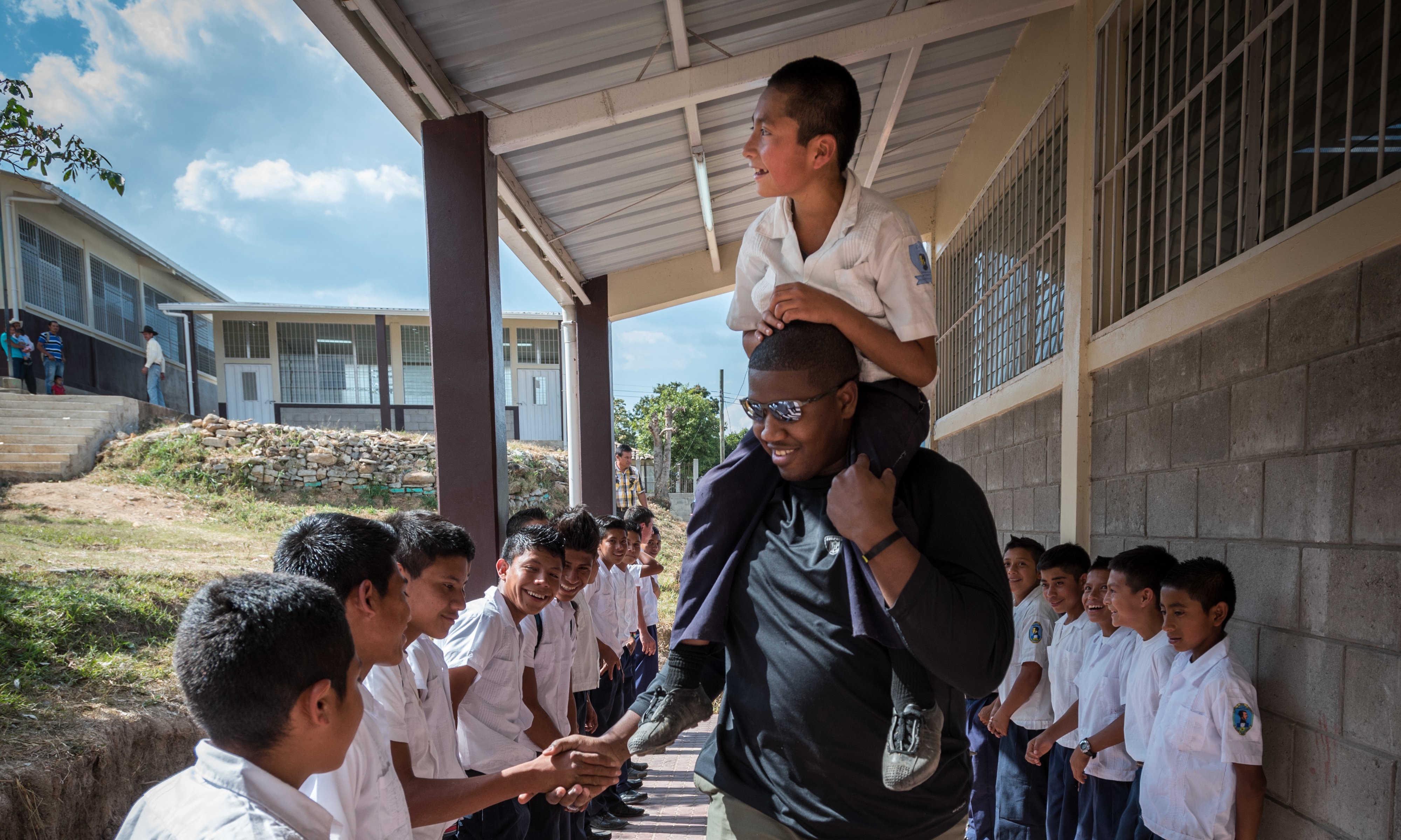 Kelvin wearing sunglasses carries a smiling boy on his shoulders while shaking hands with students lined up outside a school building.