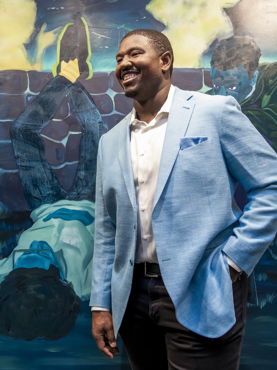 Kelvin in a light blue blazer and white shirt stands in front of a colorful mural painting.