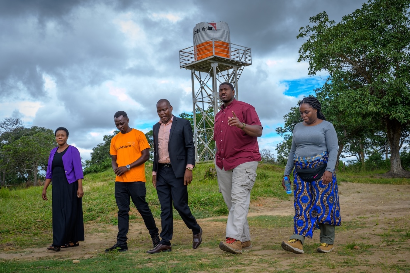 Five people walking outdoors near a water tower with 'World Vision' logo under a cloudy sky.