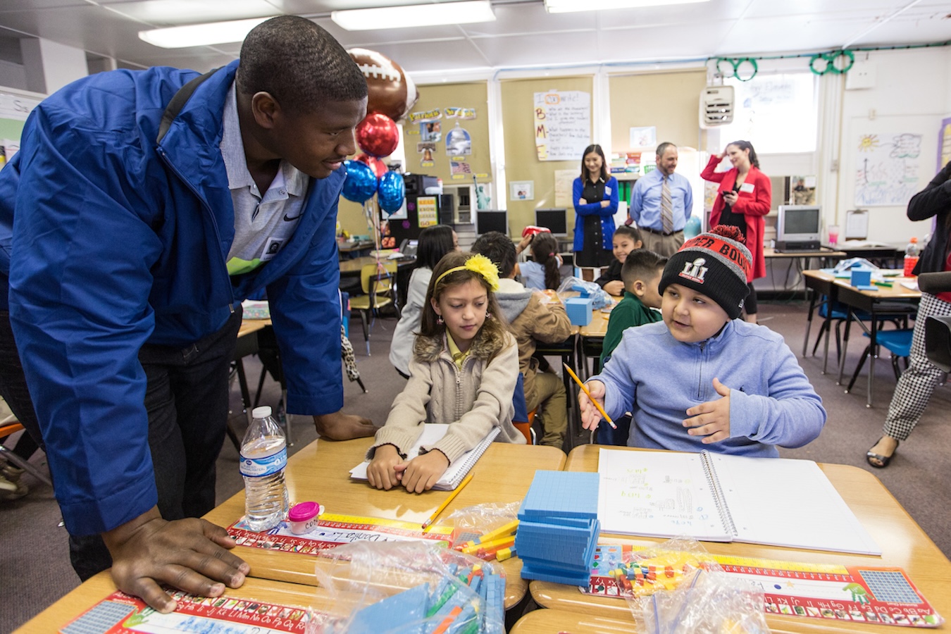 Kelvin in blue jacket leaning over to assist two elementary students sitting at a desk with school supplies in a classroom.