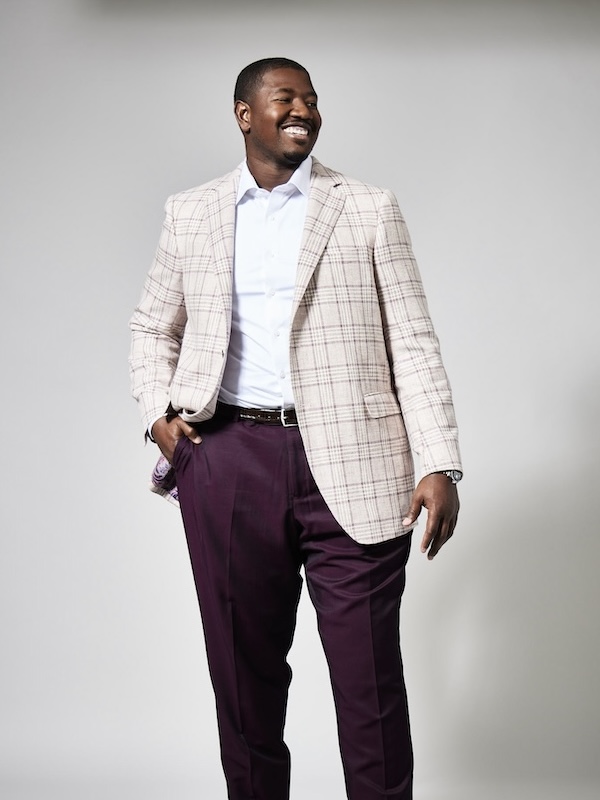 Smiling man wearing a light plaid blazer, white shirt, and dark purple pants against a plain gray background.