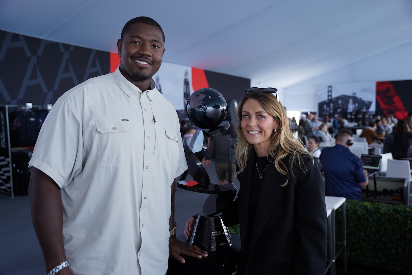 Kelvin in a white shirt and a woman in a black jacket pose beside a black trophy with a globe on top inside a tent with people and event displays in the background.