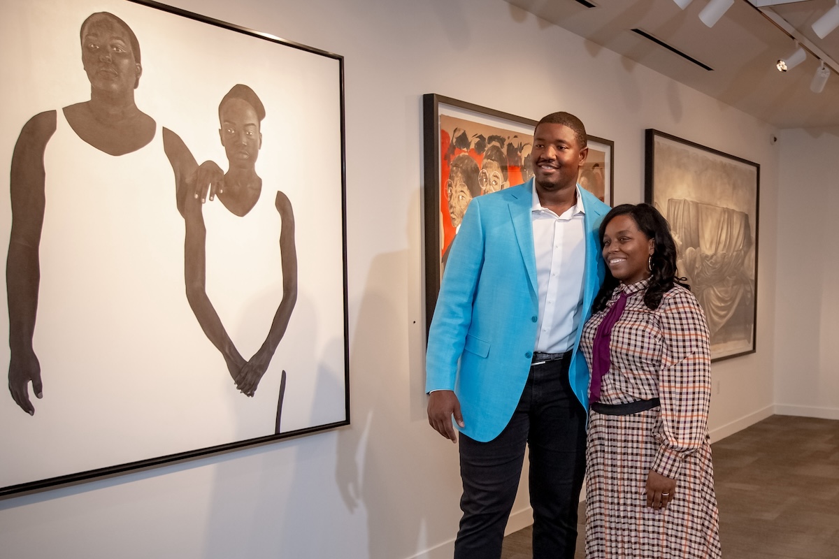 Kelvin in a light blue blazer and a woman in a checkered dress smiling and posing together in an art gallery with framed paintings on the wall.