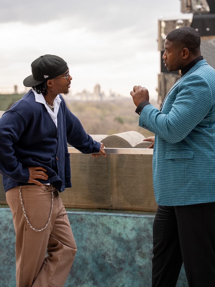 Kelvin engaged in a conversation on a balcony overlooking a blurred cityscape and trees, one wearing a blue jacket and tan pants with a chain, the other in a blue checked blazer and black pants.