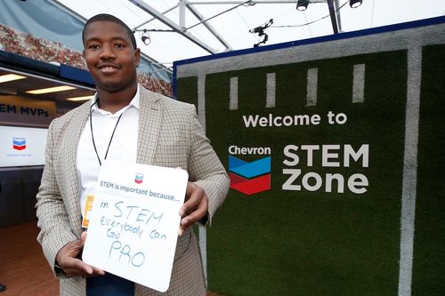 Kelvin holding a whiteboard that says 'in STEM everybody can go pro' standing next to a Chevron STEM Zone sign on green turf.