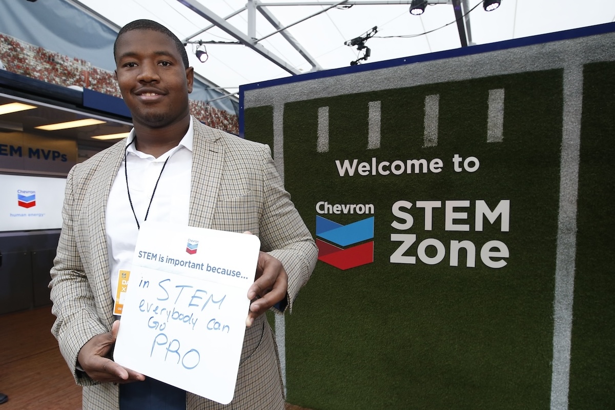 Kelvin holding a whiteboard that says 'in STEM everybody can go pro' standing next to a Chevron STEM Zone sign on green turf.