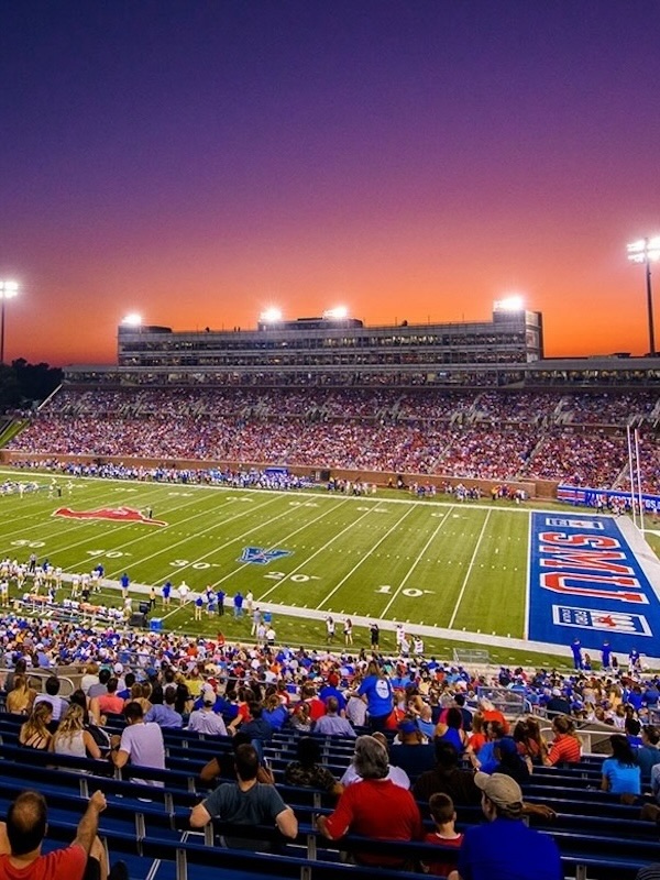Crowded football stadium at sunset with fans watching a game on a field marked with SMU Mustangs logo and team colors.