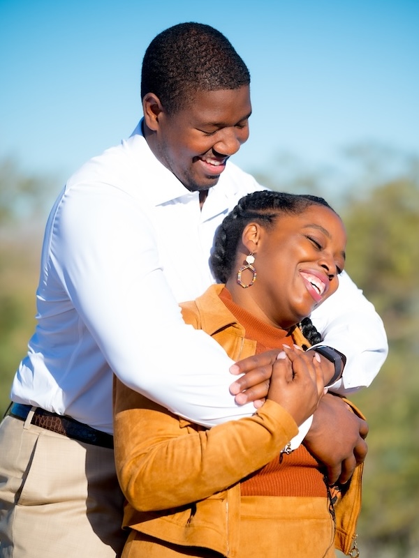 Kelvin in white shirt hugging his laughing wife Jessica in brown jacket outdoors.