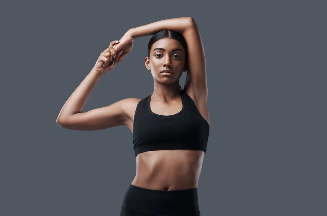 Schwarz gekleidetes weibliches Fitnessmodel dehnt sich vor einem grauen Hintergrund in einer Holmes Place-Trainingsumgebung.