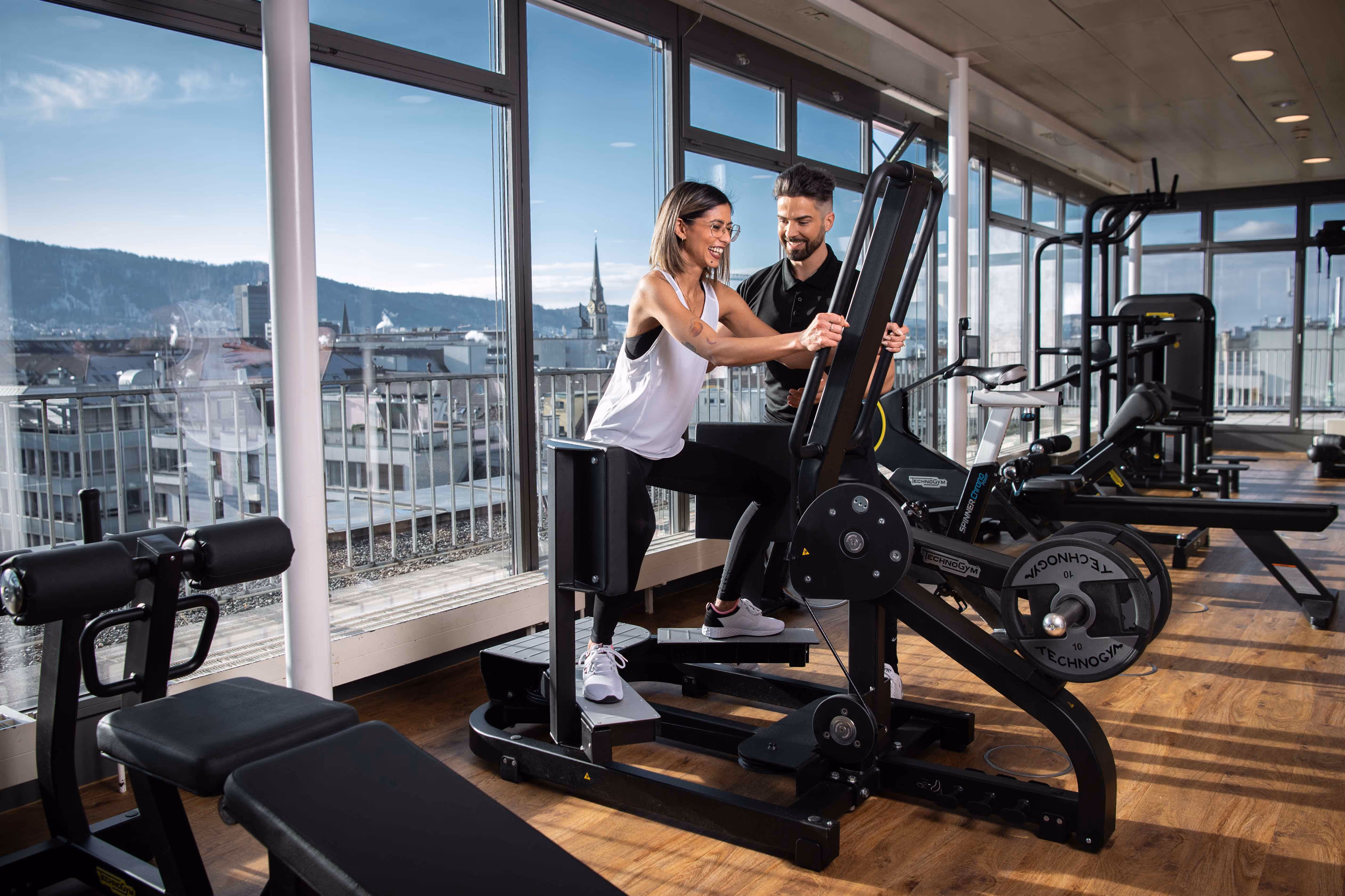 Frau trainiert unter Anleitung eines Personal Trainers an Fitnessgeräten von Holmes Place mit Blick auf die Stadt.