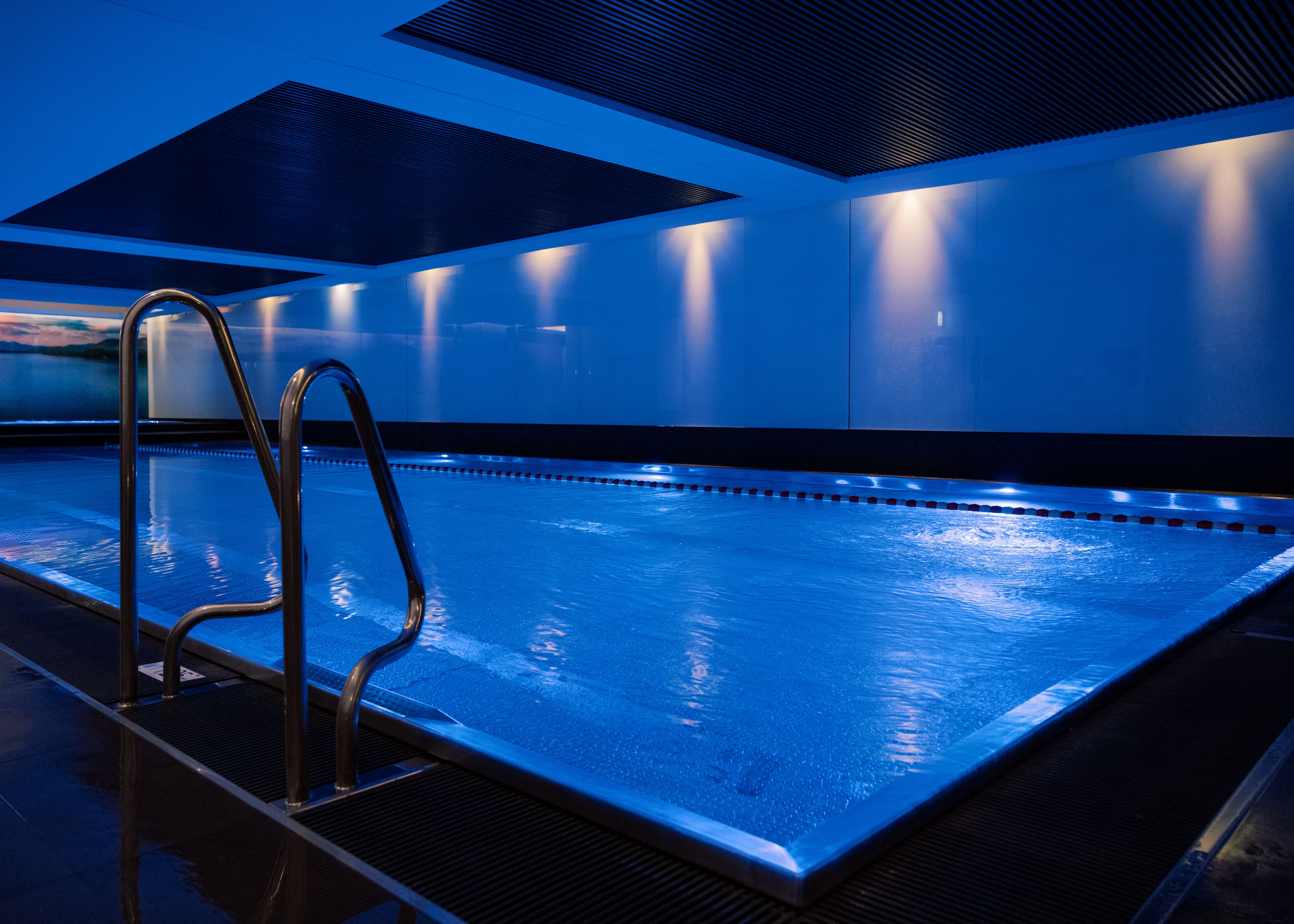 Schwimmen Sie im Innenpool von Holmes Place mit blauer Beleuchtung und einer eleganten Metallleiter.