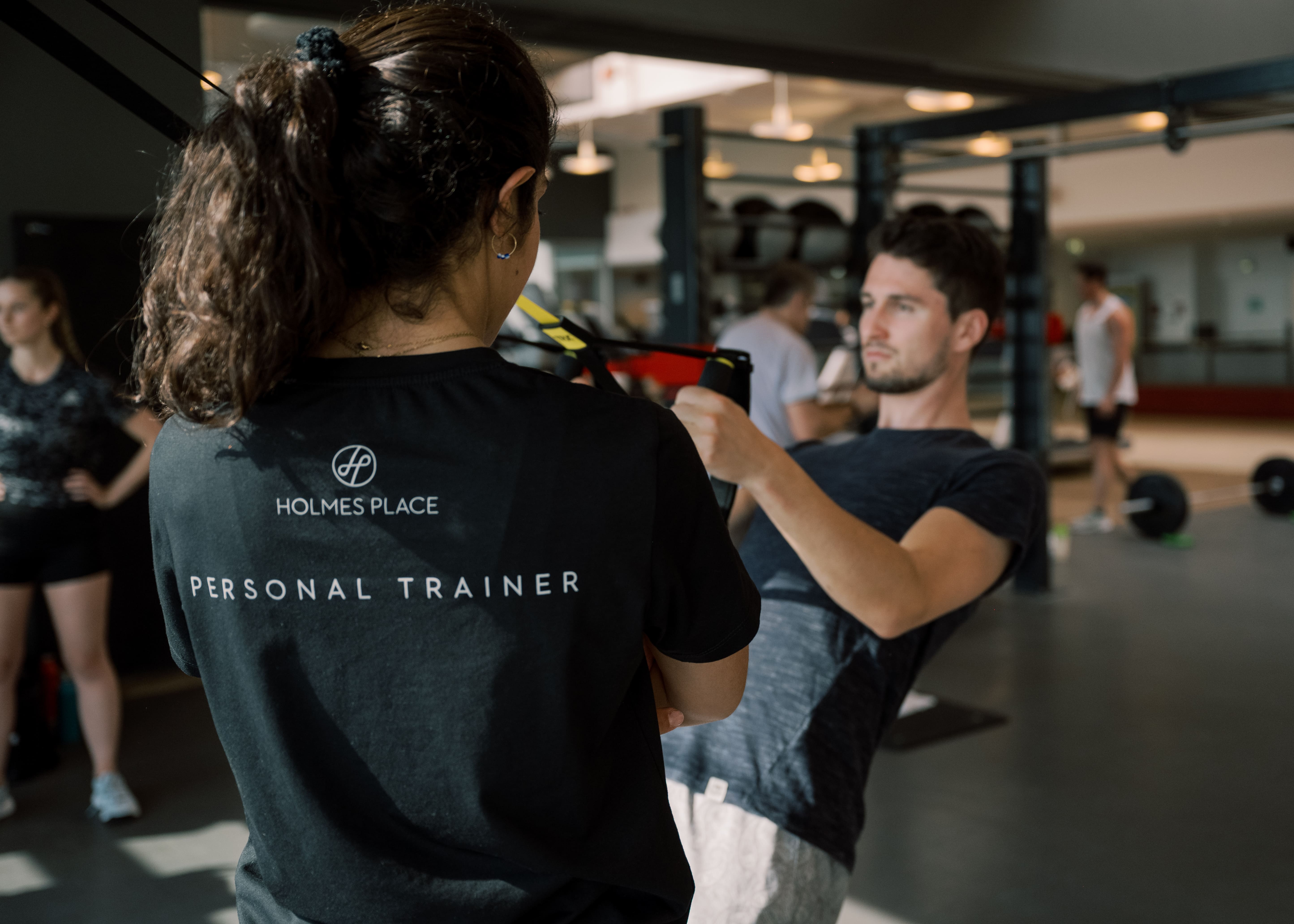 Der Personal Trainer von Holmes Place führt seinen Kunden unter fachkundiger Aufsicht durch die Übungen in einem modernen Fitnessstudio.