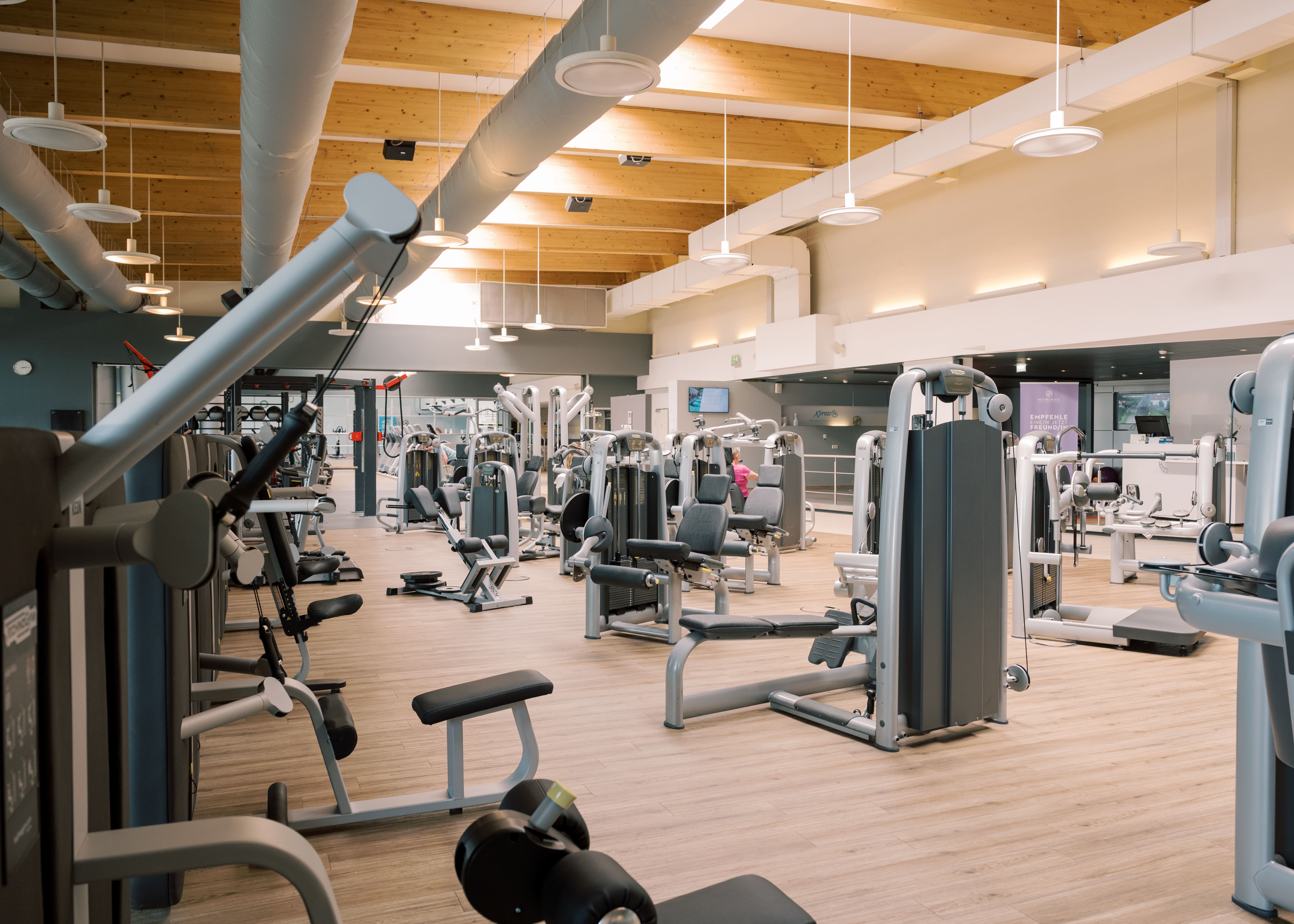 Inneneinrichtung des Fitnessstudios von Holmes Place mit moderner Beleuchtung und Trainingsgeräten auf Holzbalken.