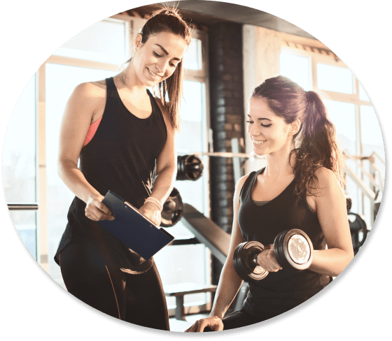 A fitness trainer with a clipboard coaching a woman lifting a dumbbell in a gym.
