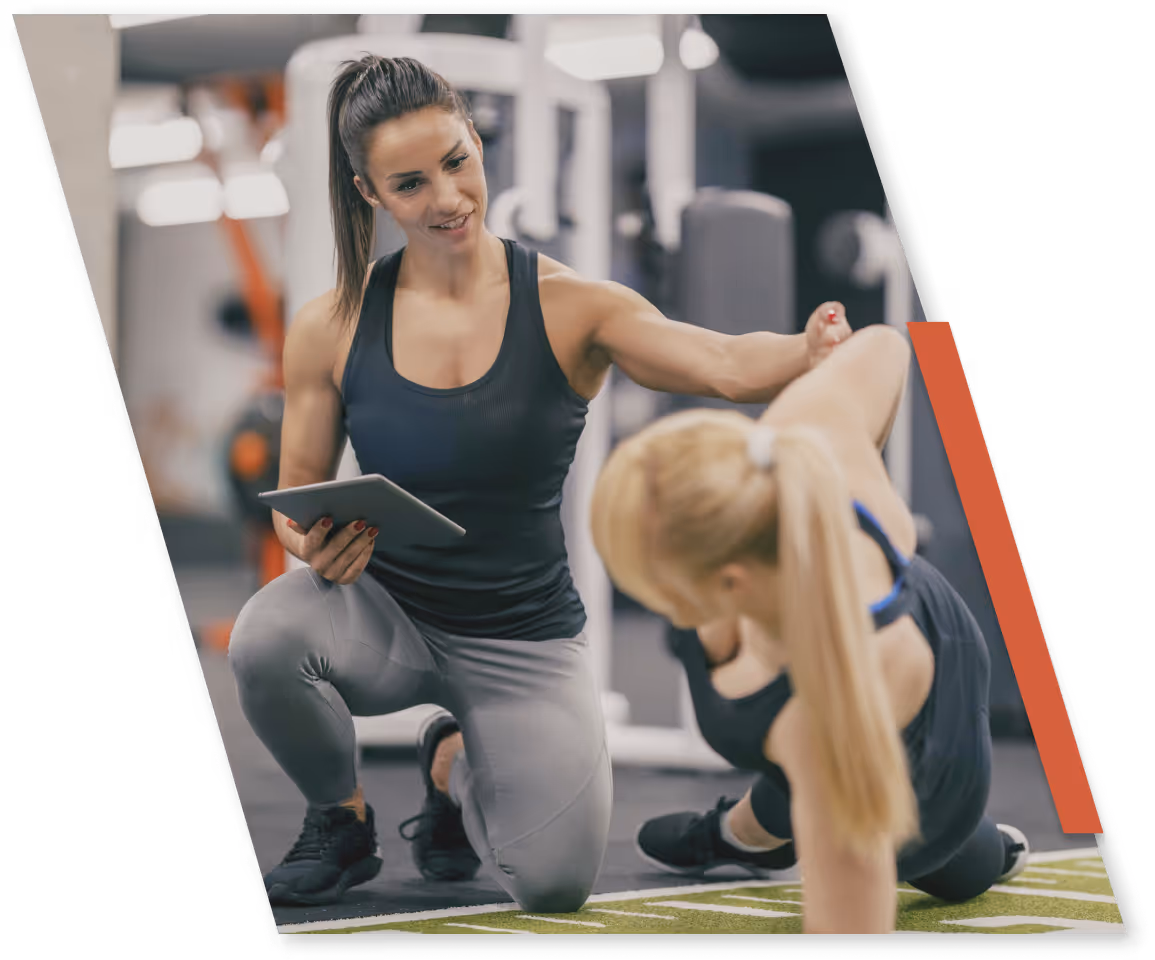Female fitness coach kneeling and holding a tablet while guiding a woman exercising on a mat in a gym.