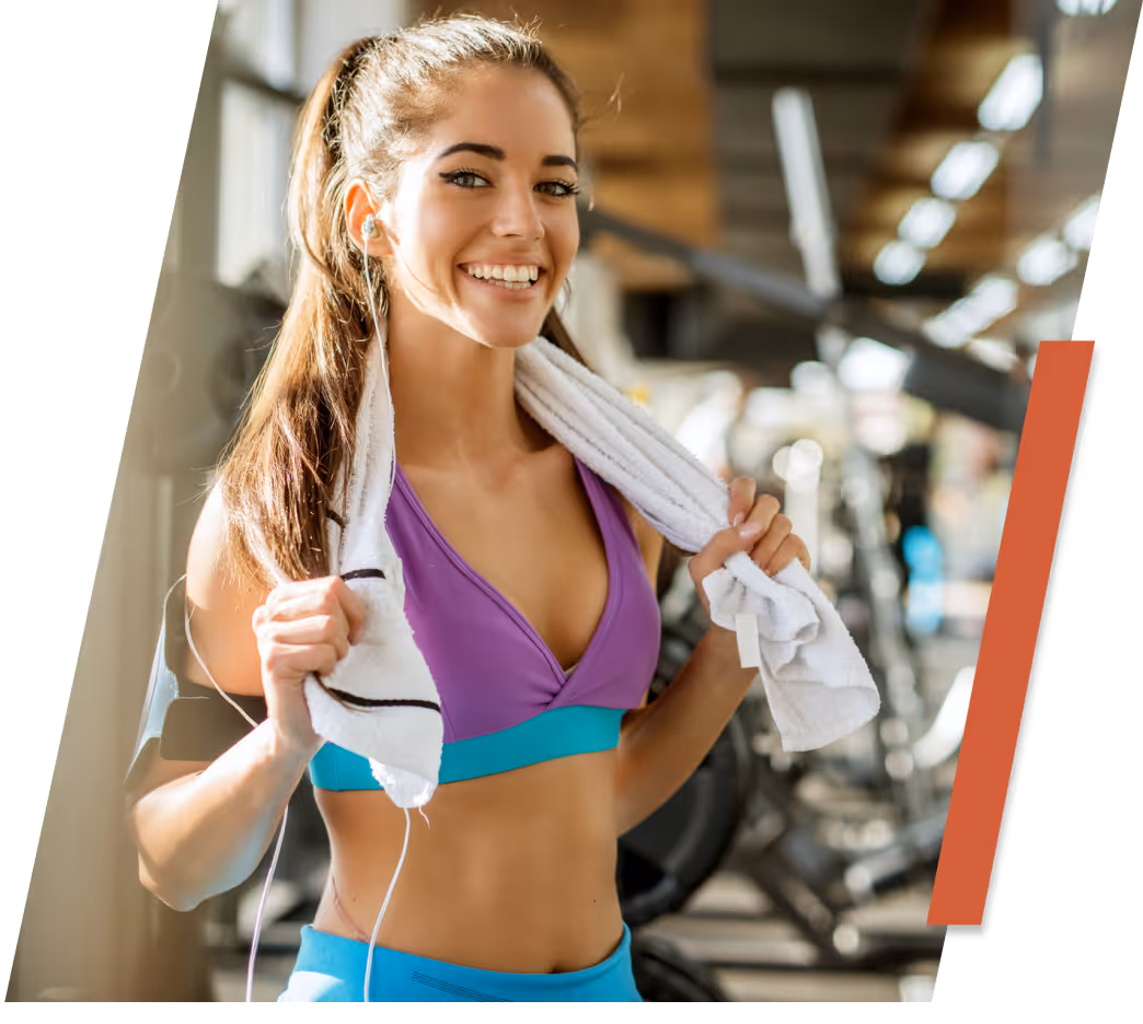Smiling young woman in workout clothes holding a white towel around her neck at a gym.