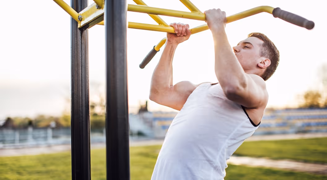 Man in a white sleeveless shirt performing pull-ups on a yellow outdoor exercise bar.