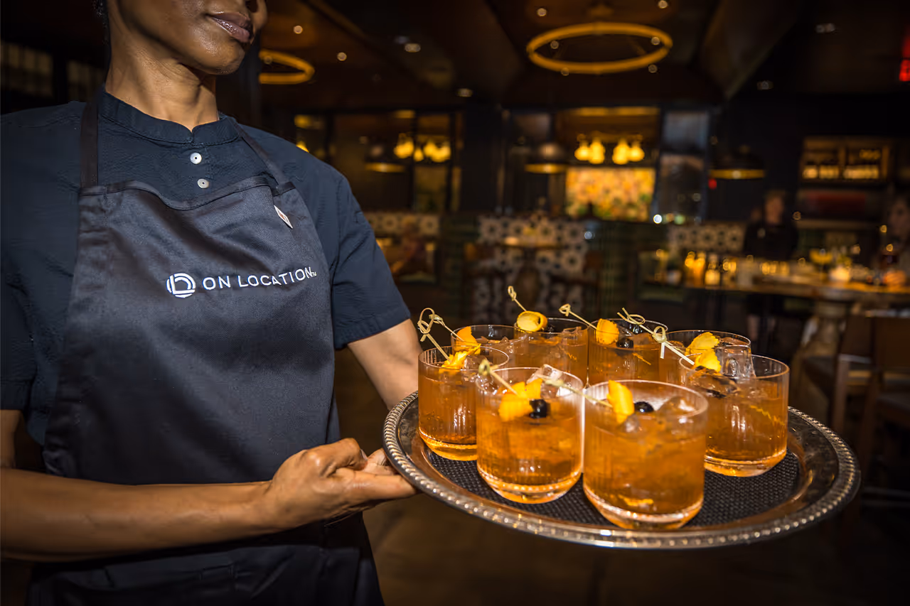 Server carrying tray of craft cocktails with orange garnishes in dimly lit bar