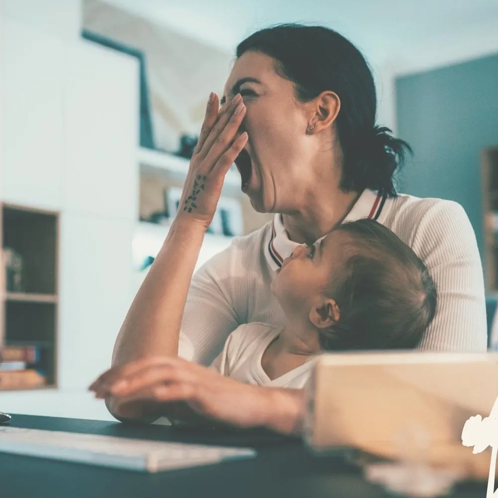 Eine Frau gähnt und hält ein Kleinkind auf dem Arm, während sie an einem Schreibtisch sitzt.