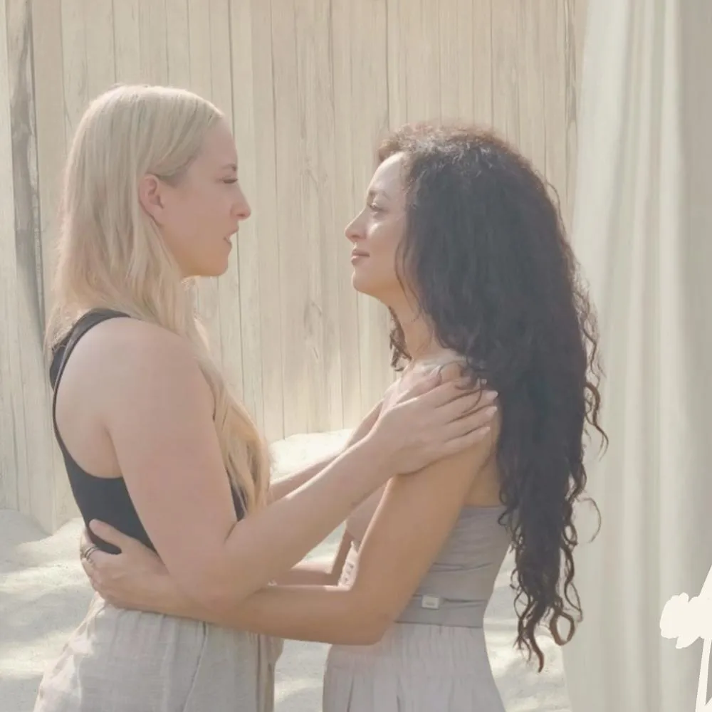 Zwei Frauen umarmen und blicken sich liebevoll in die Augen vor einem hellen Holzhintergrund.