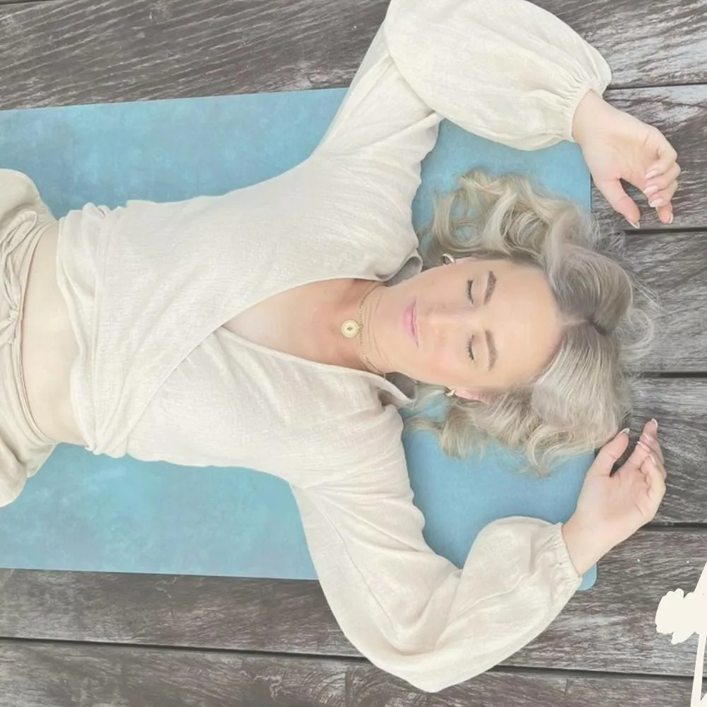 Frau mit geschlossenen Augen liegt auf einer blauen Matte auf Holzdielen und entspannt sich.