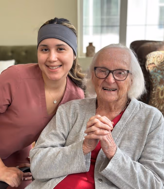 Smiling elderly woman with glasses seated wearing a red top and gray cardigan, with a young woman in a pink uniform and gray headband leaning beside her.