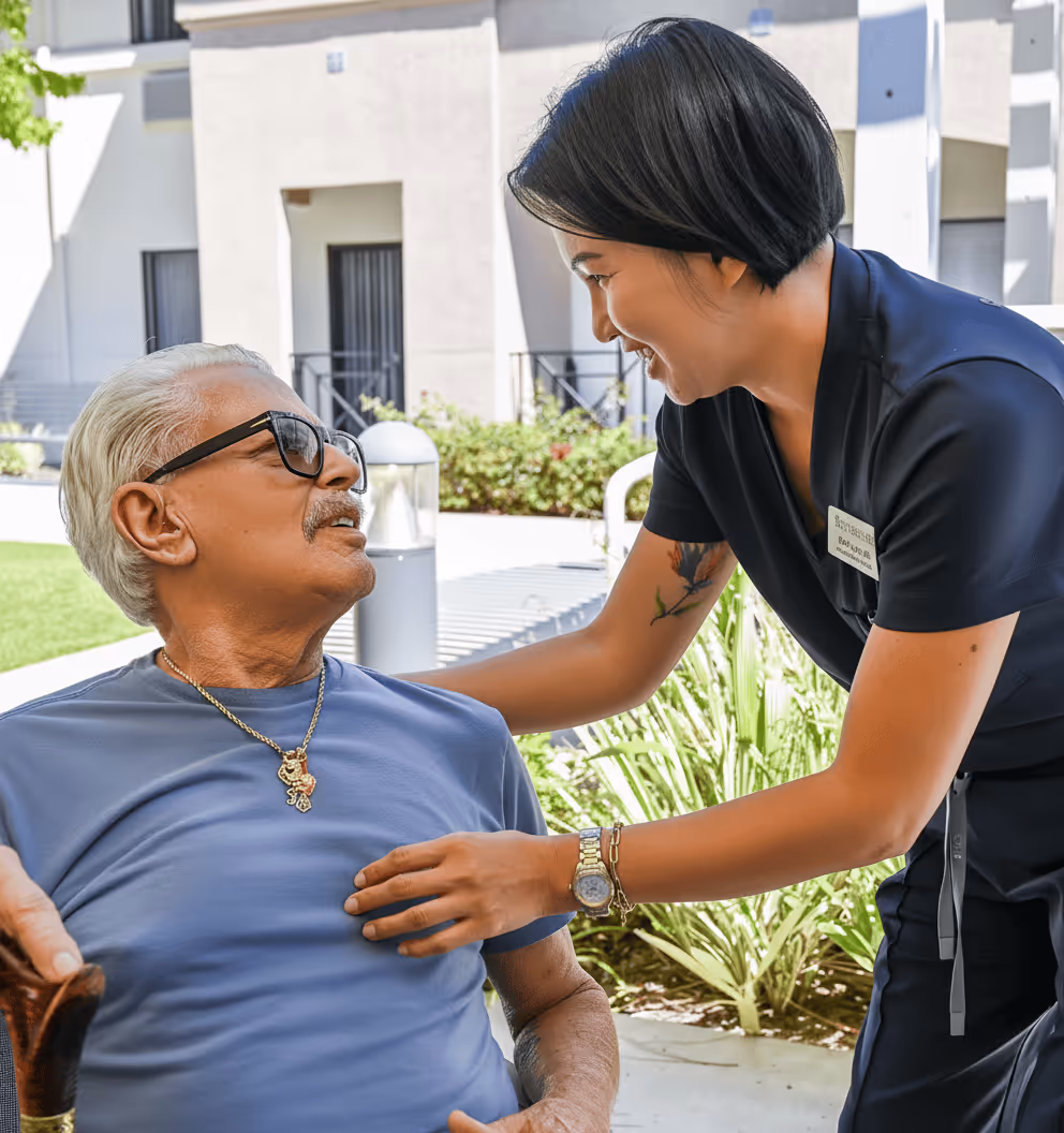 Female caregiver smiling and gently touching the arm of a seated elderly man outdoors by a building.