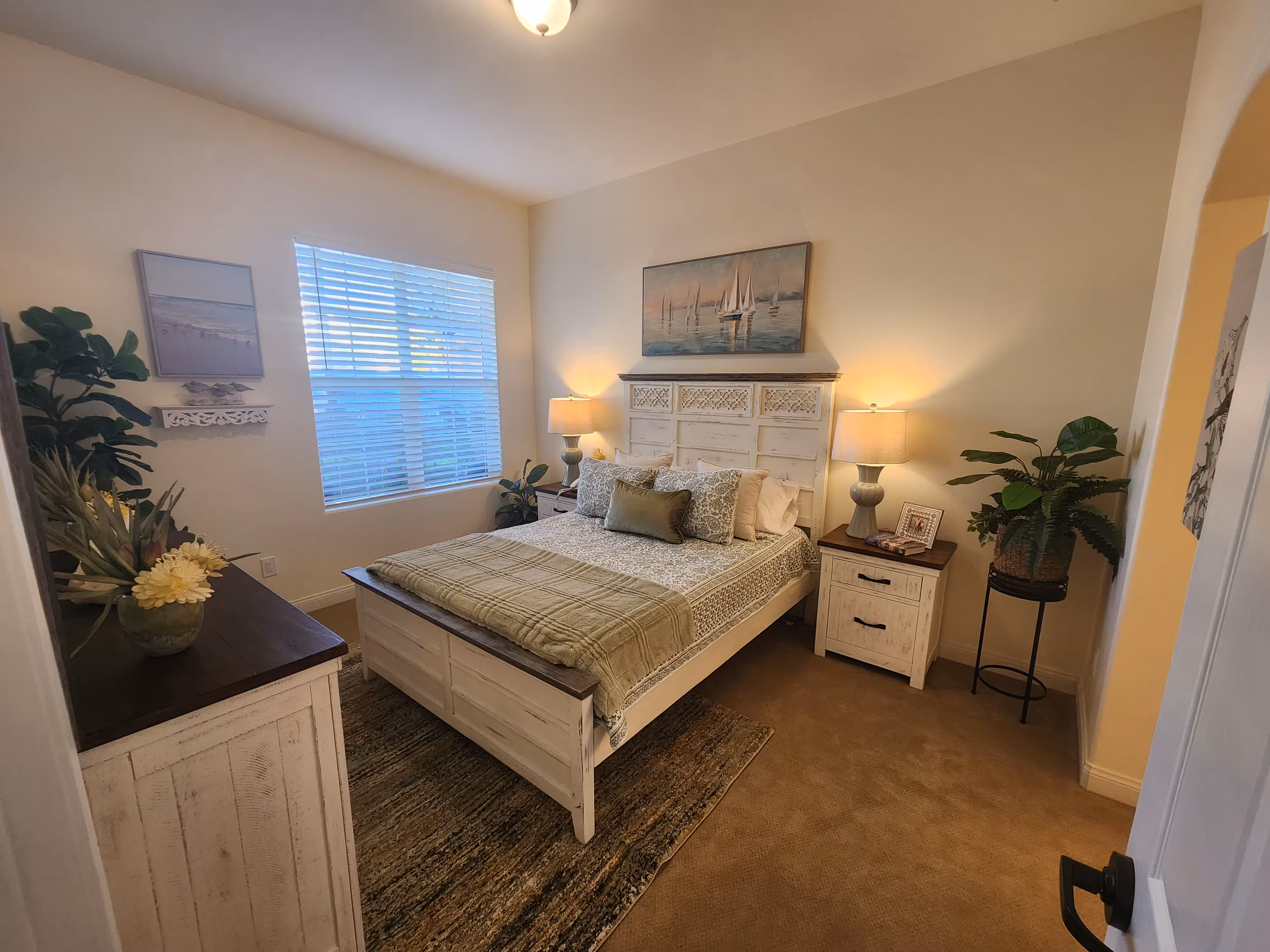 Cozy bedroom with a white wood bed, patterned bedspread, two bedside tables with lamps, plants, and a window with blinds.