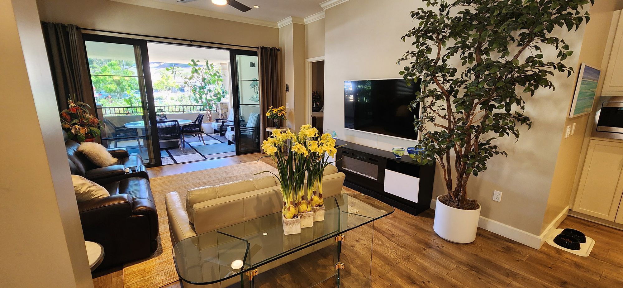 Modern living room with leather and fabric sofas, a glass table with yellow flowers, a large potted plant, wall-mounted TV, and sliding glass doors opening to a patio with outdoor seating.