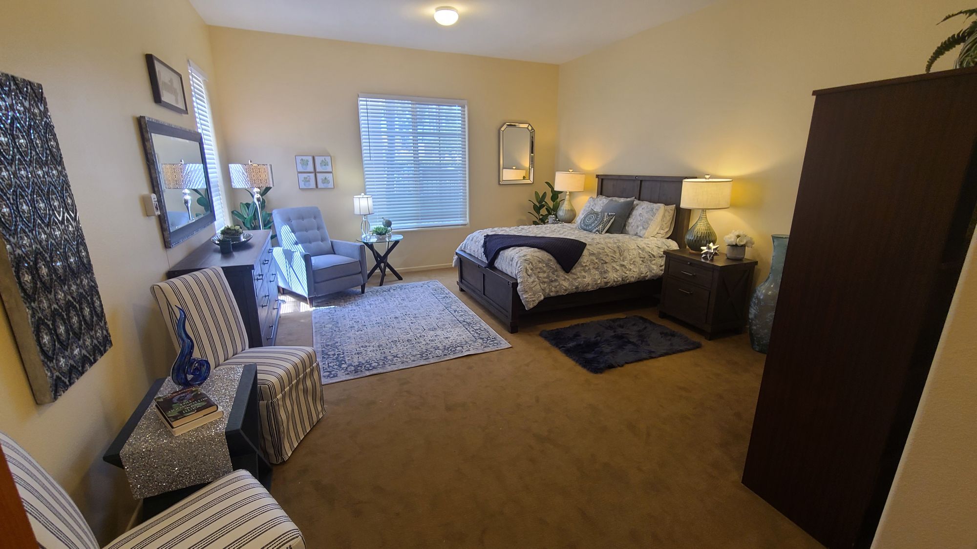 Cozy bedroom with a dark wood bed, matching nightstands, seating area with striped and blue chairs, and decorative art on beige walls.