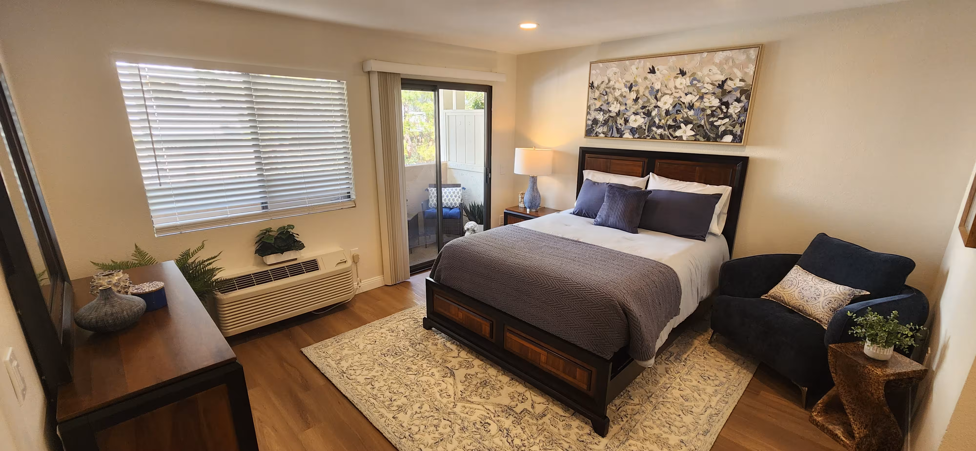 Cozy bedroom with a wooden bed, gray bedding, floral painting above, blue armchair, bedside lamp, and sliding glass door to a balcony.