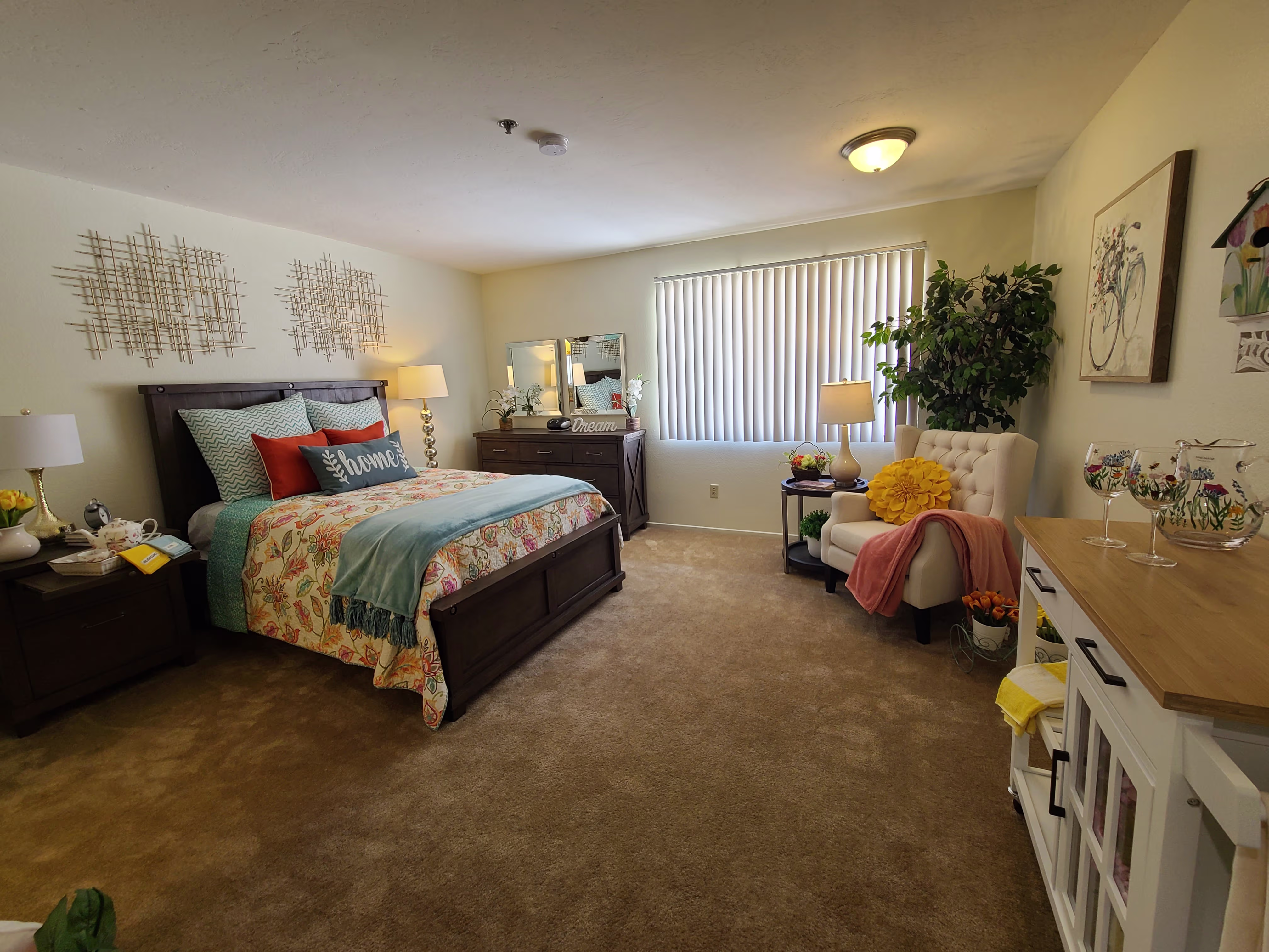 Cozy bedroom with a dark wooden bed, floral bedding, decorative pillows, nightstands, a dresser with a mirror, a cream armchair, and a window with vertical blinds.