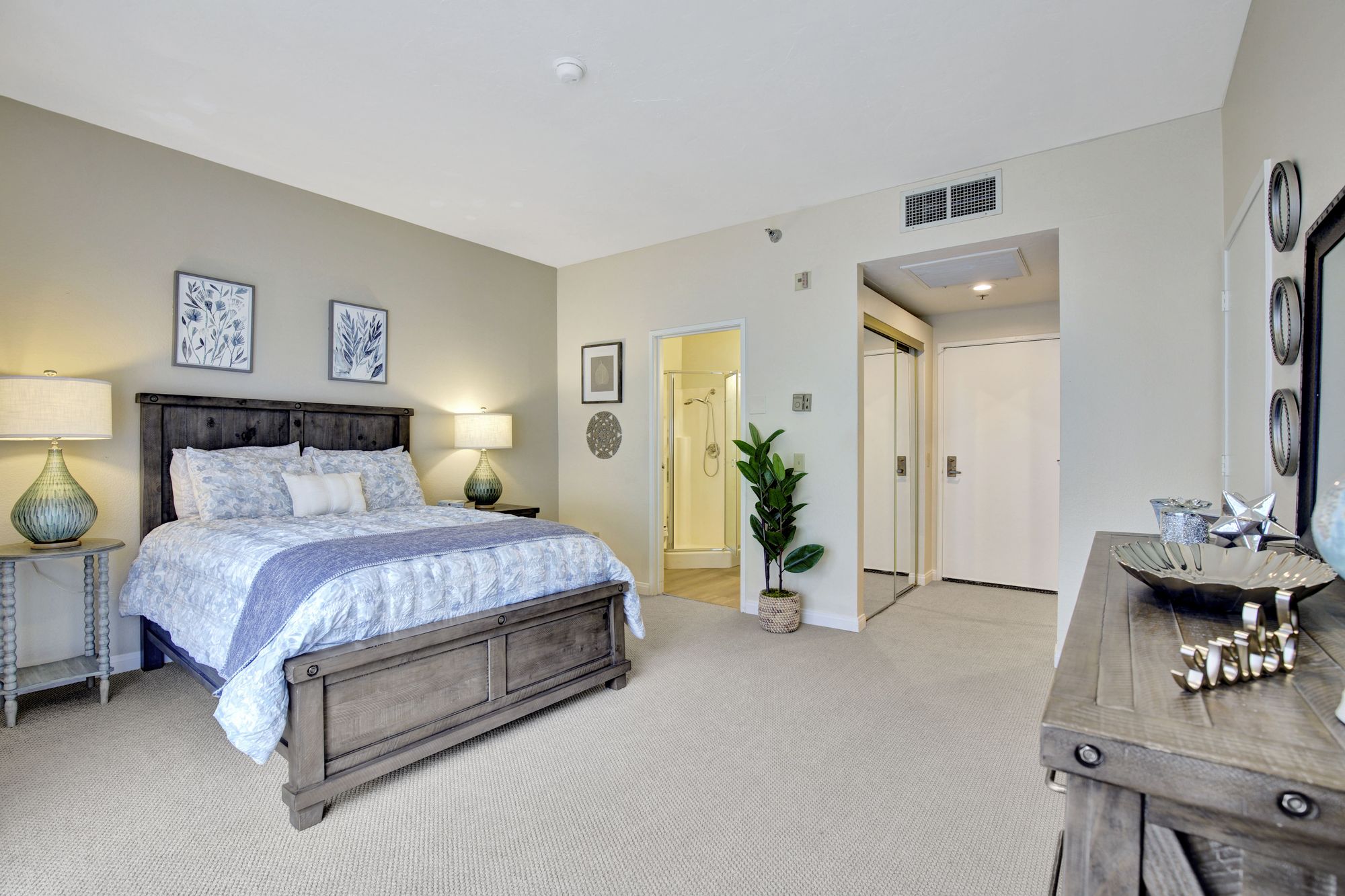 Bright bedroom with wooden queen bed, floral bedding, two lamps on side tables, a potted plant, and an open door to a bathroom with a shower.