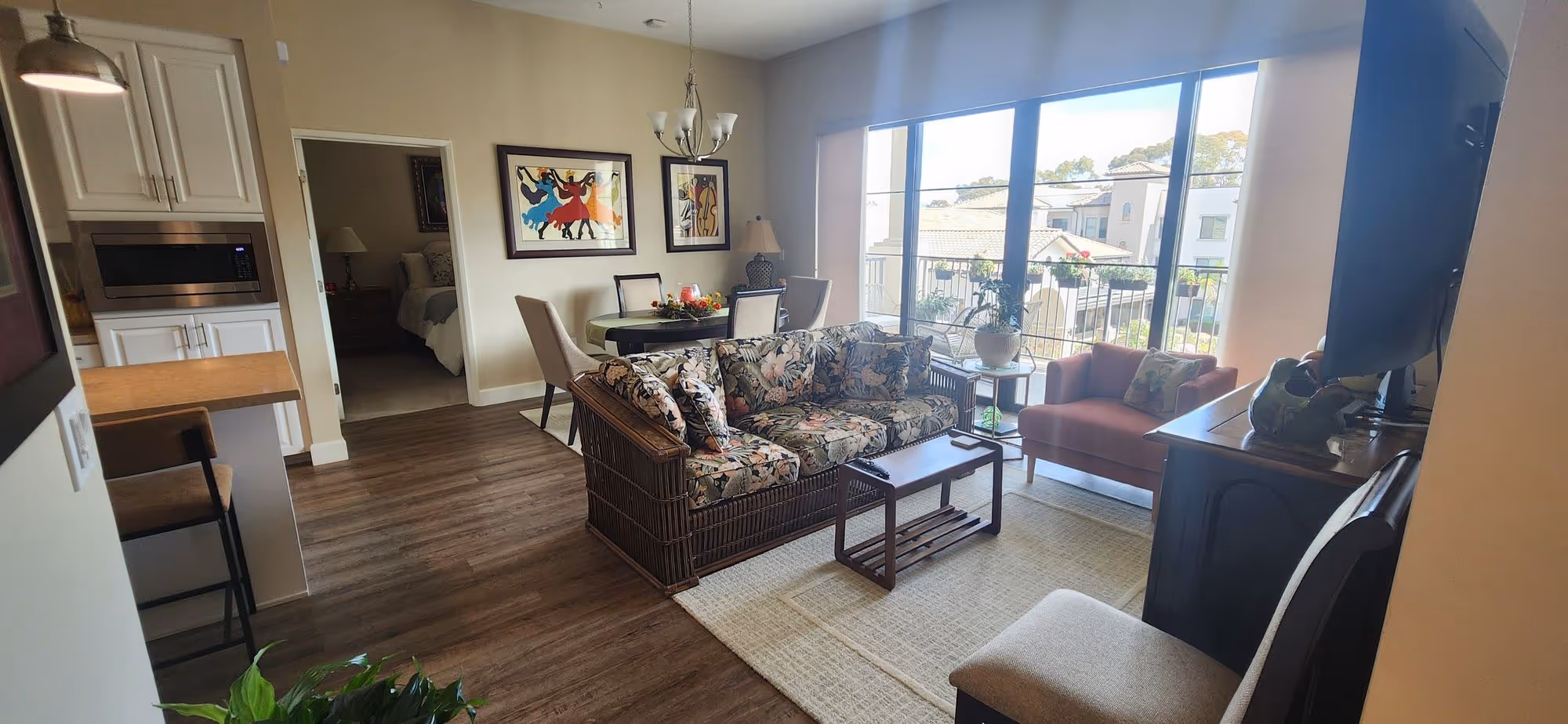 Living room with floral sofa, coffee table, armchair, dining table with chairs, and large sliding glass doors overlooking a balcony.