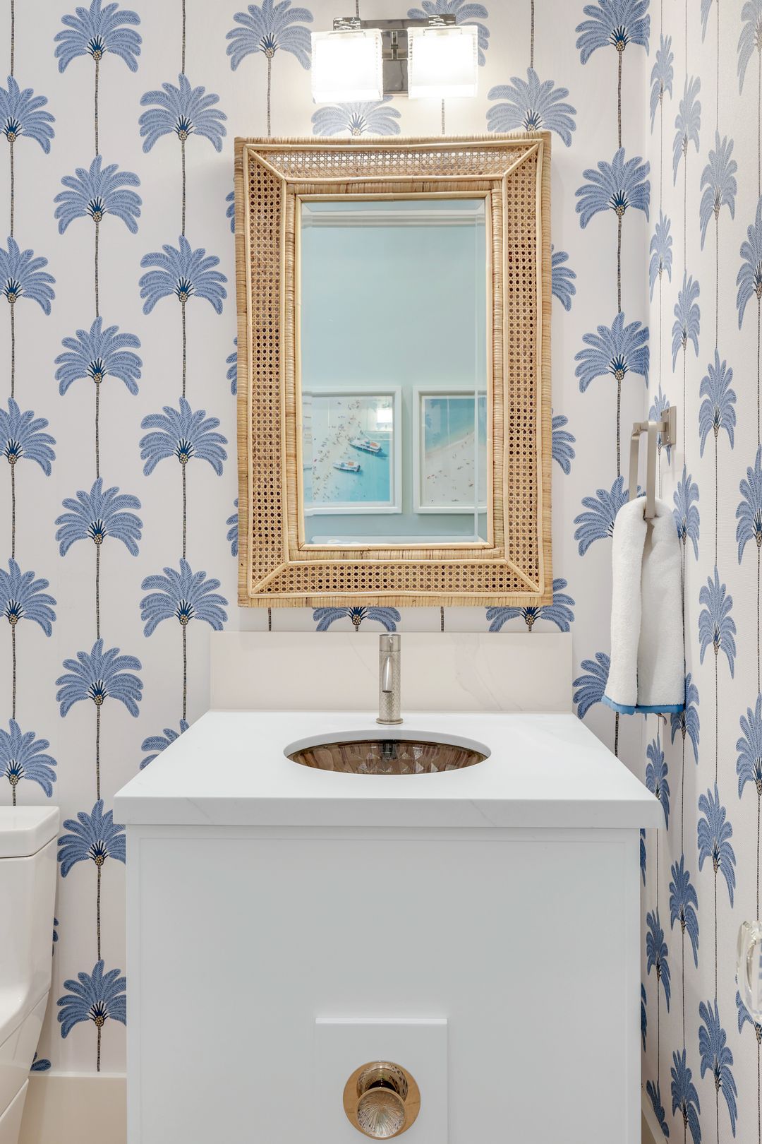 Bathroom vanity with white countertop and round sink, wicker framed mirror above, blue palm tree patterned wallpaper, and two white towels hanging on the right.