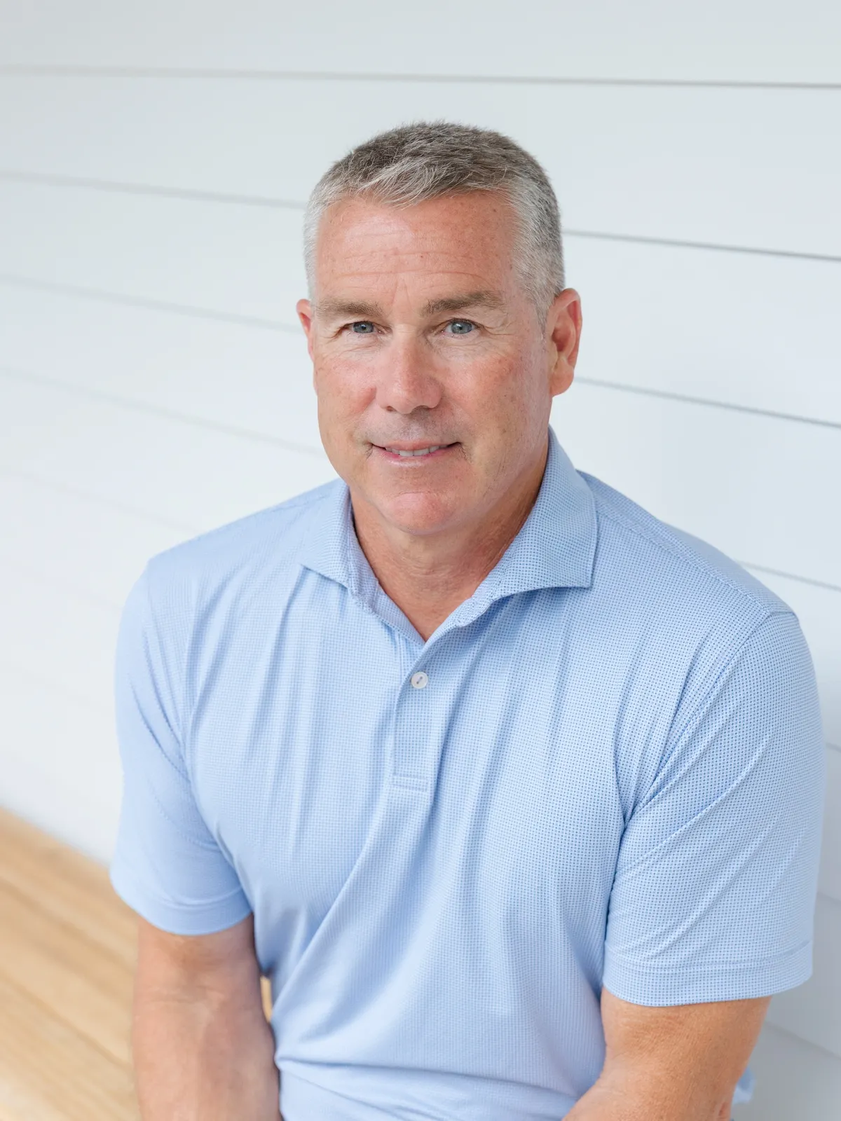 Middle-aged man with short gray hair wearing a light blue polo shirt, sitting against a white paneled wall.