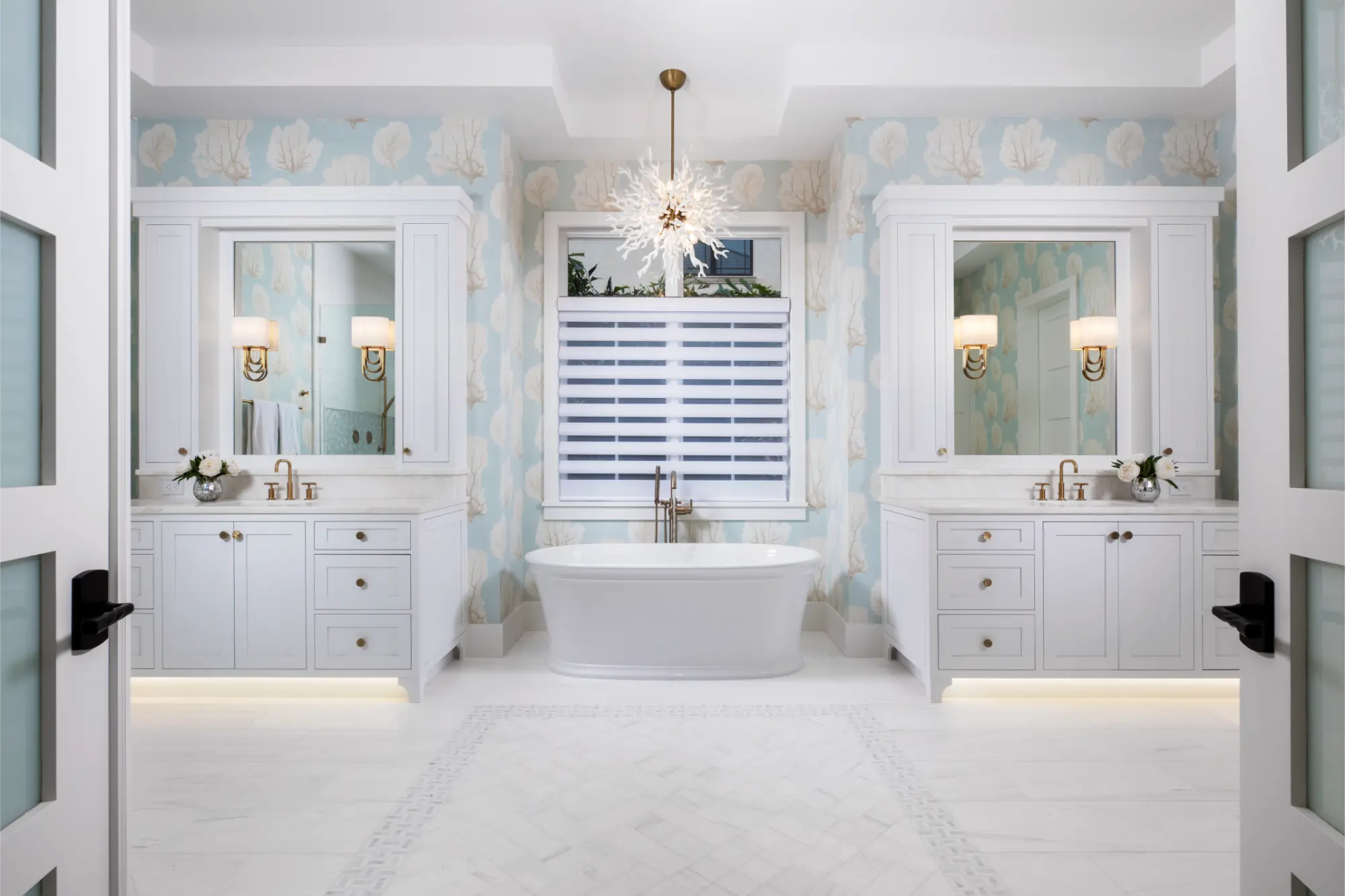 Bright bathroom with white freestanding tub centered between two white vanities with gold fixtures, floral wallpaper, and a modern chandelier.