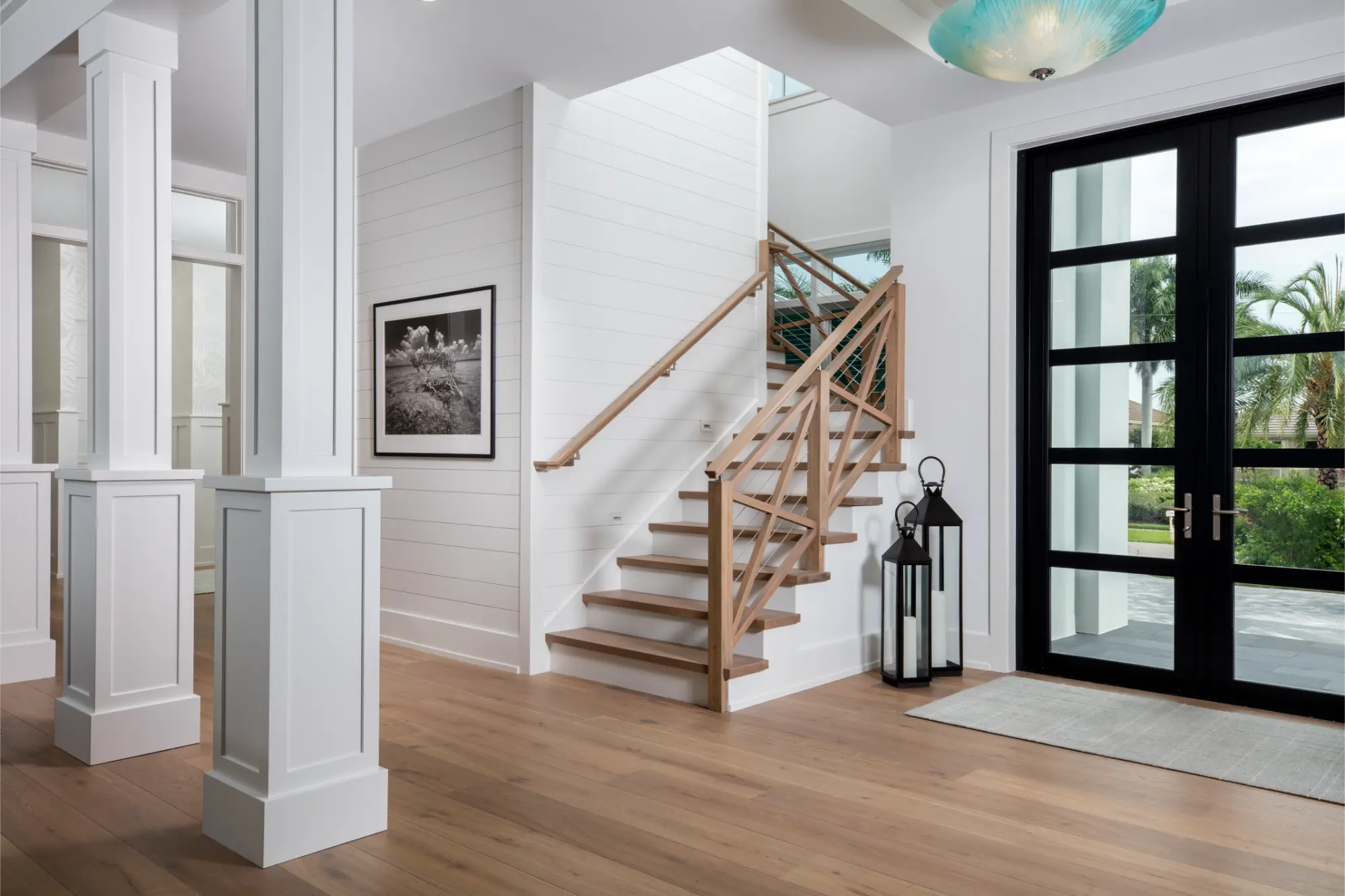 Bright entryway with wooden stairs featuring cable railing, two black lanterns, large black-framed glass doors, and white paneled columns.