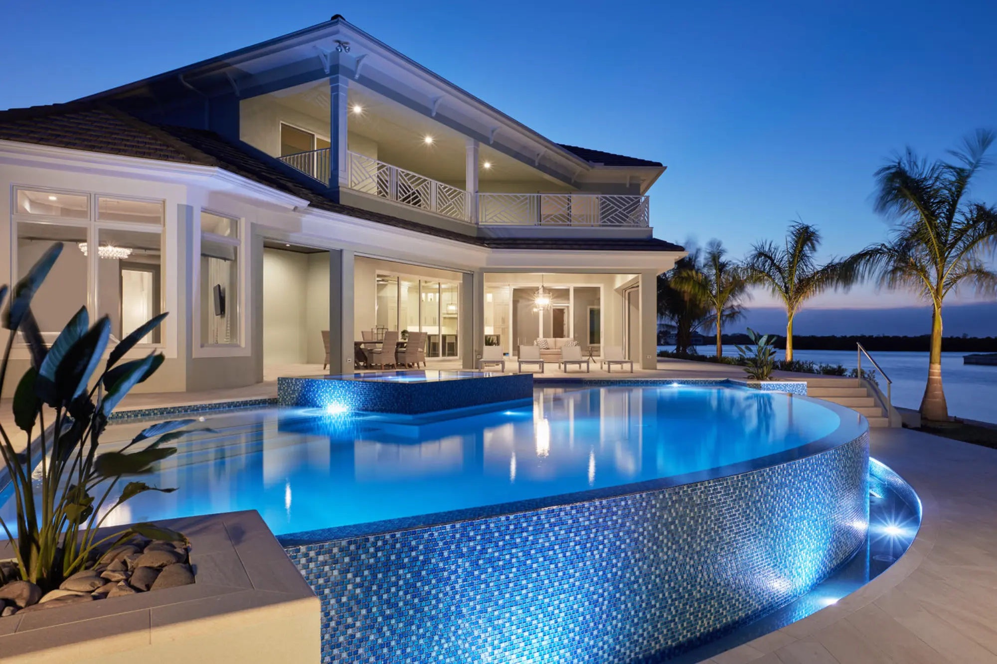 Large modern house with illuminated pool in the foreground, surrounded by palm trees at dusk.