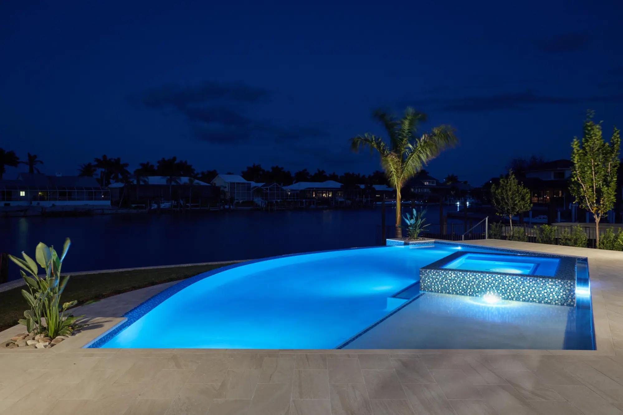 Illuminated curved swimming pool with an attached square spa at night overlooking a calm waterfront with houses and palm trees.