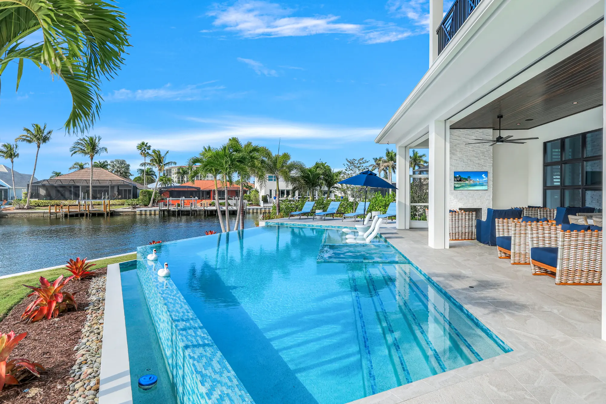 Outdoor pool area with chairs and umbrellas, overlooking a waterway and palm trees under a clear sky.
