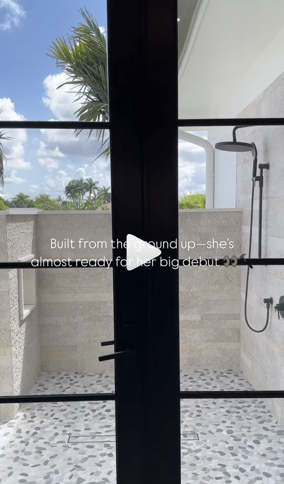 Outdoor shower area with pebble-patterned floor, beige tiled walls, and black shower fixtures, viewed through black framed glass doors with a blue sky and palm trees in the background.