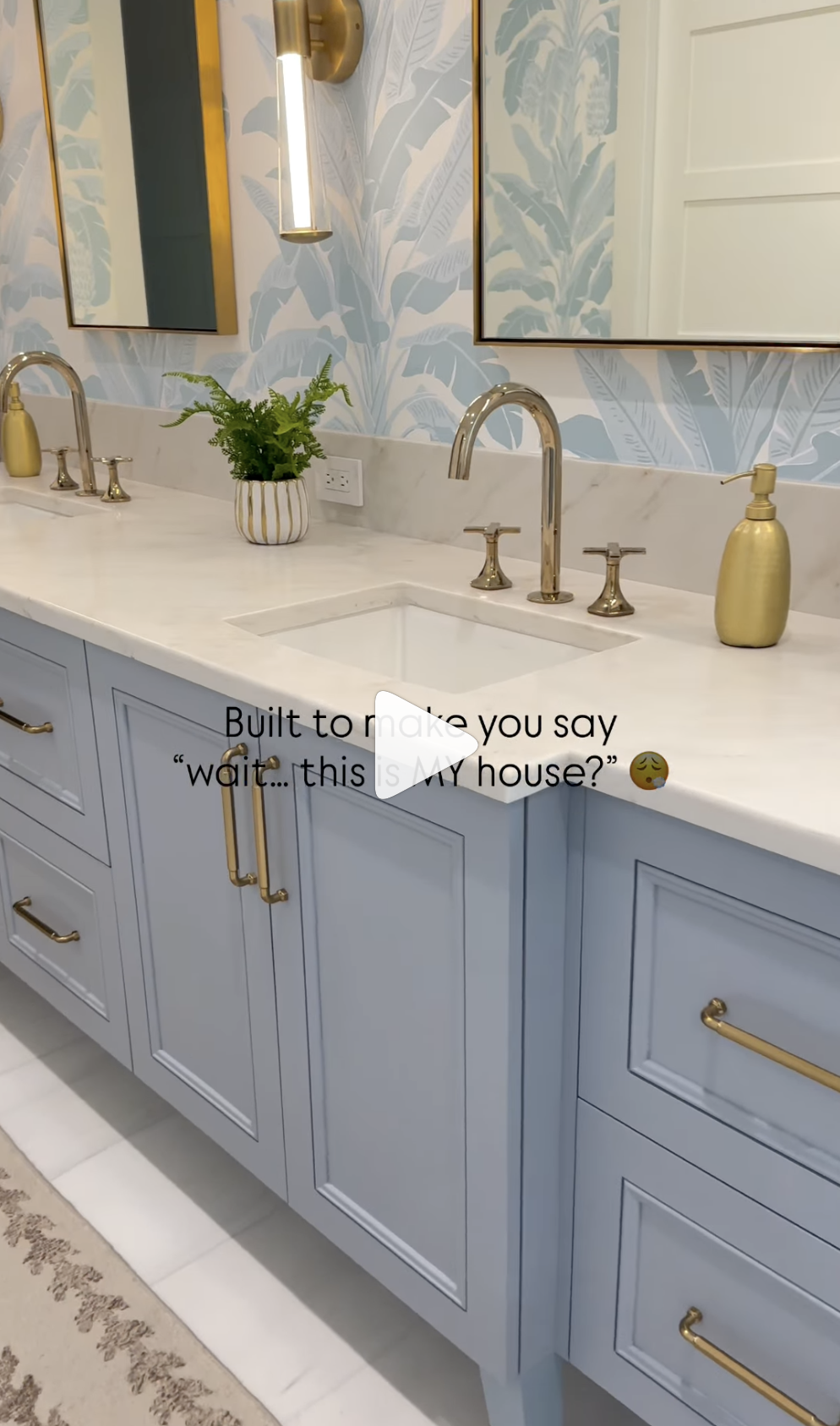 Modern bathroom vanity with light blue cabinets, gold hardware, dual sinks, and leafy wallpaper above the white marble countertop.