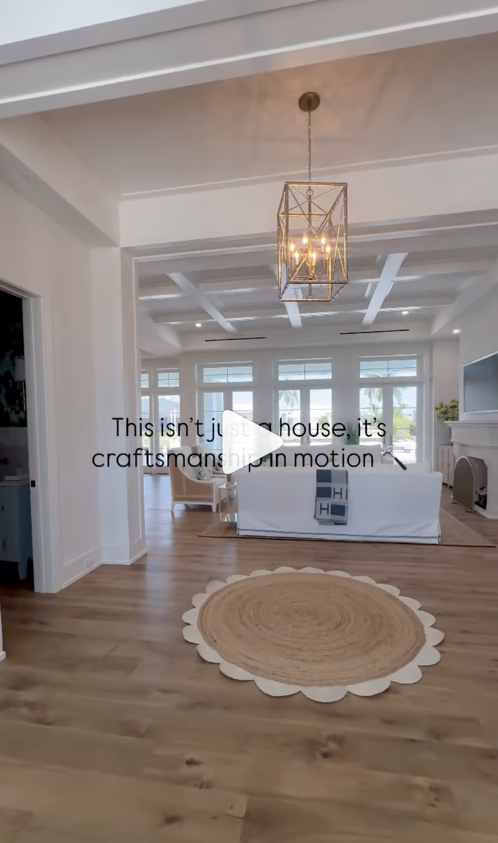 Bright living room with wood flooring, a round rug, white sofa, armchair, coffered ceiling, and large windows showing palm trees outside.
