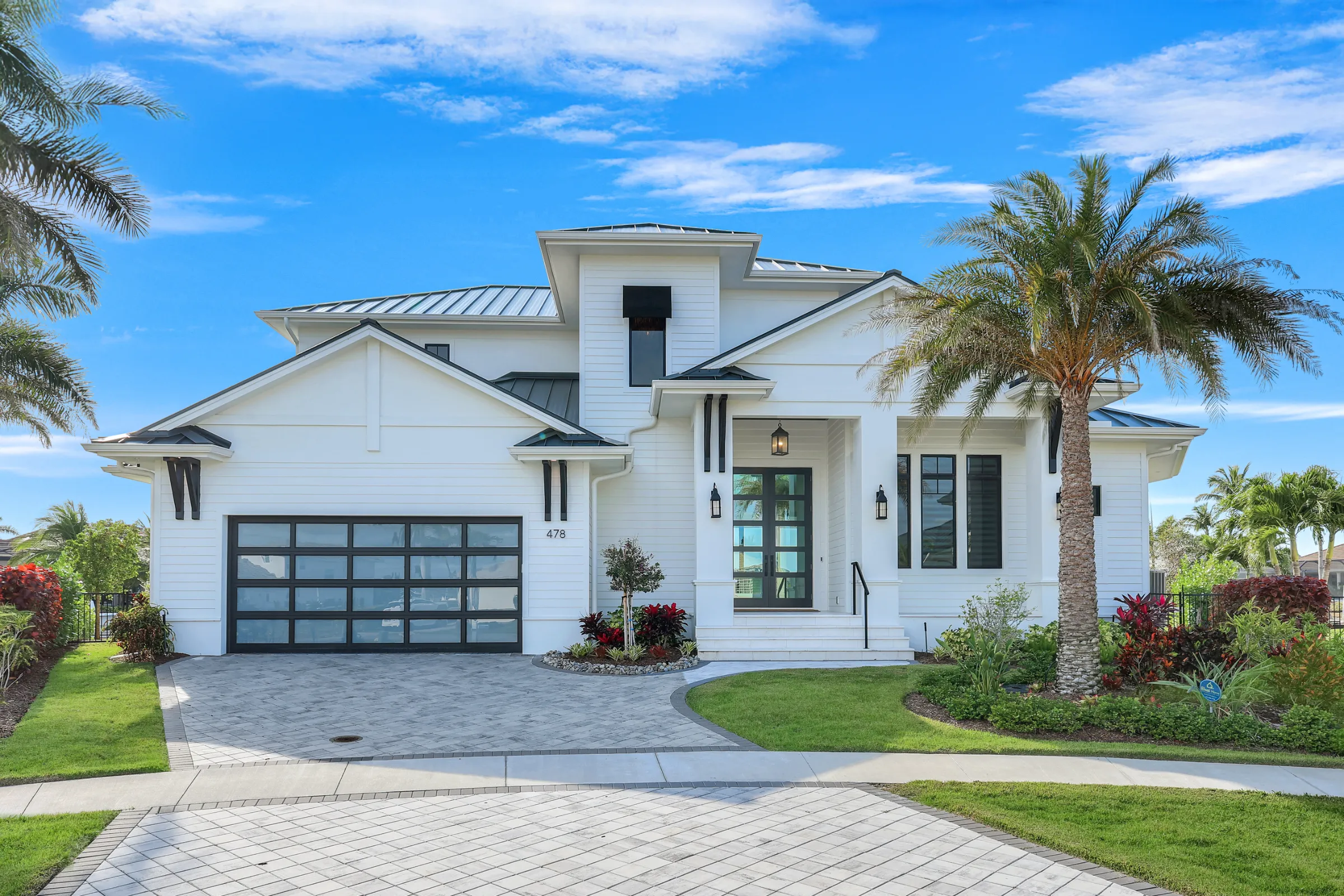Modern white two-story house with a glass-paneled garage door, landscaped front yard, and palm trees under a blue sky.