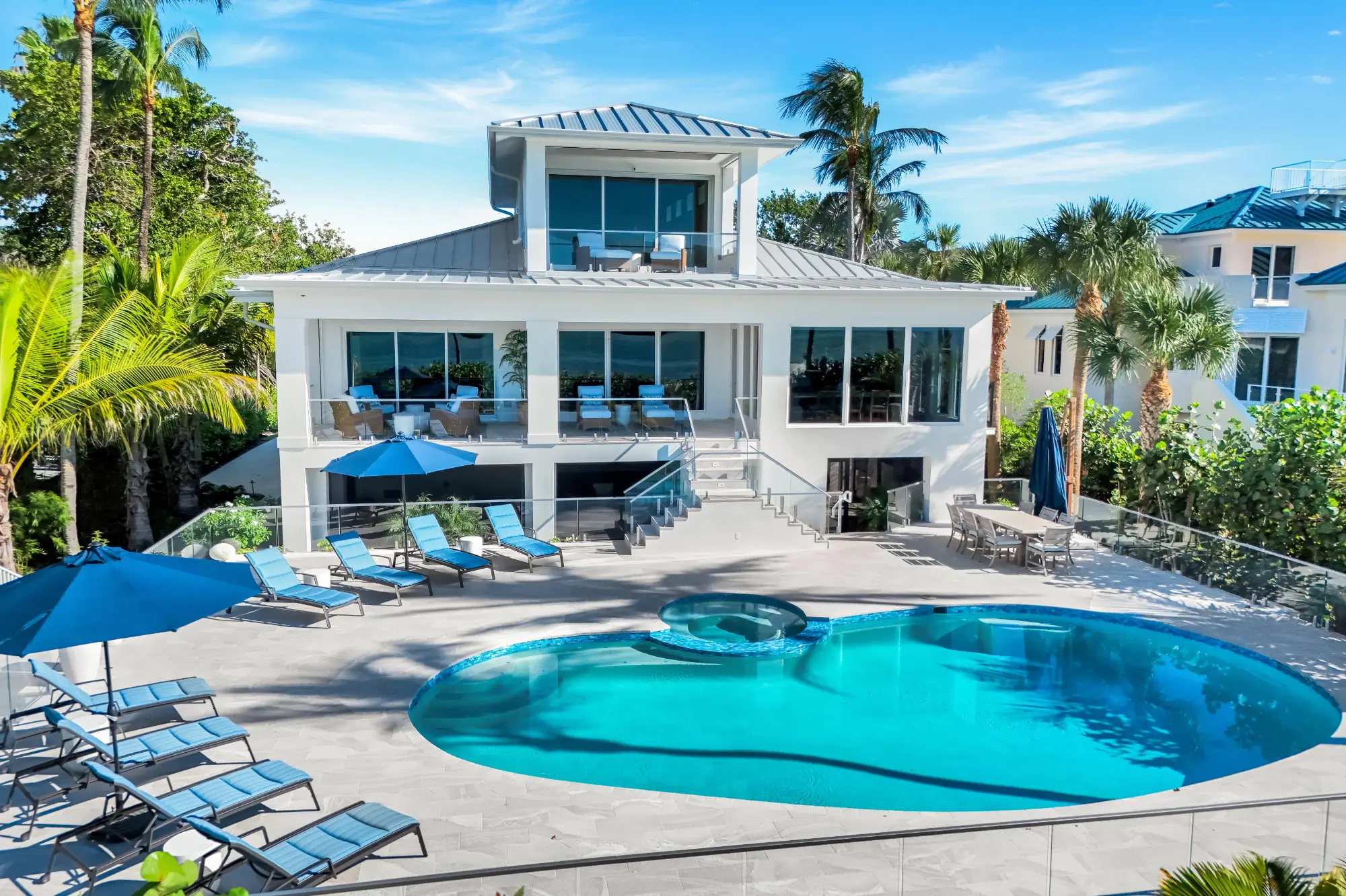 Modern white two-story house with large windows, a kidney-shaped swimming pool, blue lounge chairs, umbrellas, and palm trees under a clear blue sky.